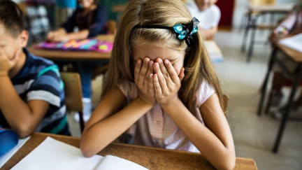 Little girl in the classroom feeling stressed and covering her face with hands while sitting at the desk.