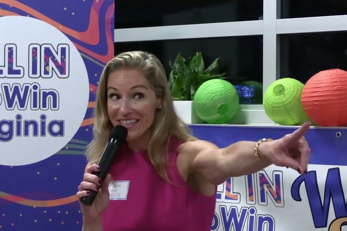 A blonde woman (Susanna Gibson) speaks excitedly into a microphone and points broadly during a campaign event.