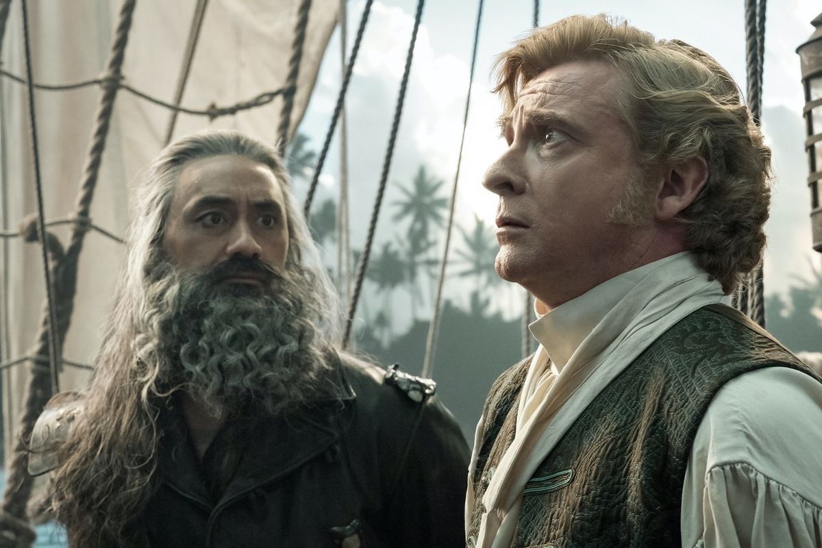 Taika Waititi and Rhys Darby in a scene from Max's 'Our Flag Means Death.' They are standing on a sailing ship in period clothing from the 1800s. Waititi is a Pacific Islander with brown skin, long salt-and-pepper hair, and a long, curly salt-and-pepper beard wearing all black. Darby is a white man with short, curly and coiffed blondish-brownish hair and thick sideburns. Waititi is looking at Darby as Darby is looking up at something worriedly.