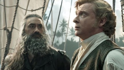 Taika Waititi and Rhys Darby in a scene from Max's 'Our Flag Means Death.' They are standing on a sailing ship in period clothing from the 1800s. Waititi is a Pacific Islander with brown skin, long salt-and-pepper hair, and a long, curly salt-and-pepper beard wearing all black. Darby is a white man with short, curly and coiffed blondish-brownish hair and thick sideburns. Waititi is looking at Darby as Darby is looking up at something worriedly.