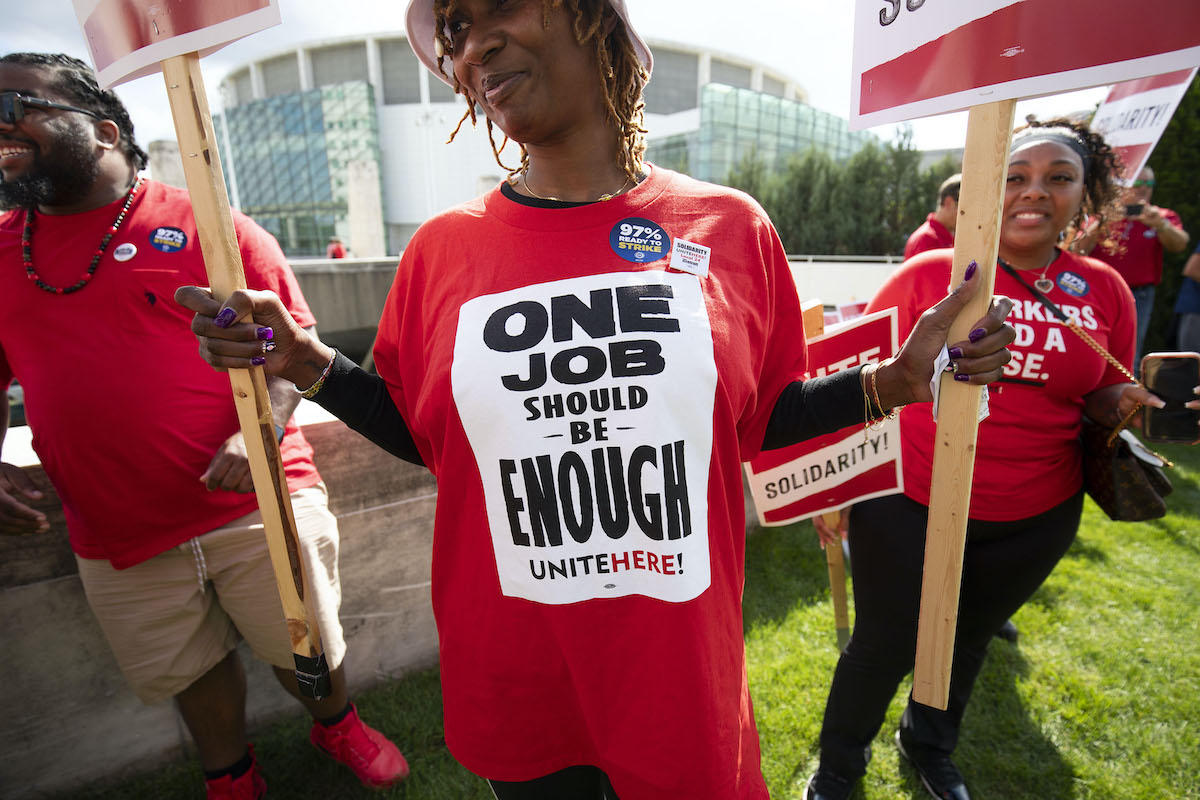 Striking workers holding picket signs. One Black woman wears a shirt reading "One job should be enough"