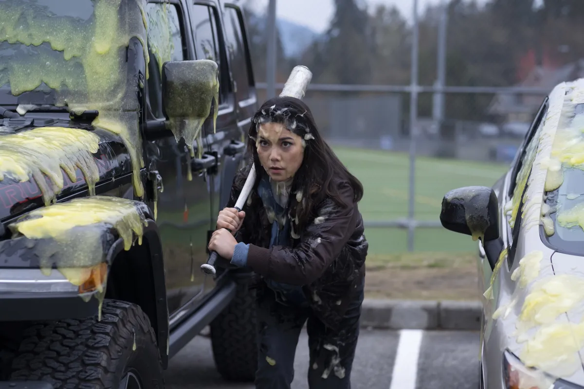 A teen girl covered in slime holding a baseball bat hides behind an SUV in a parking lot.