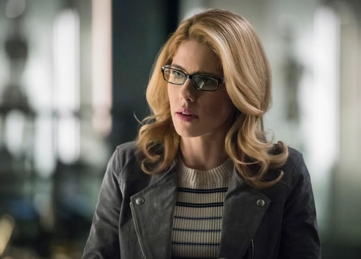 Image of Emily Bett Rickards as Felicity on The CW's 'Arrow.' She is a young woman with long blonde hair wearing black rimmed glasses, a black and white striped sweater and a grey jacket. 