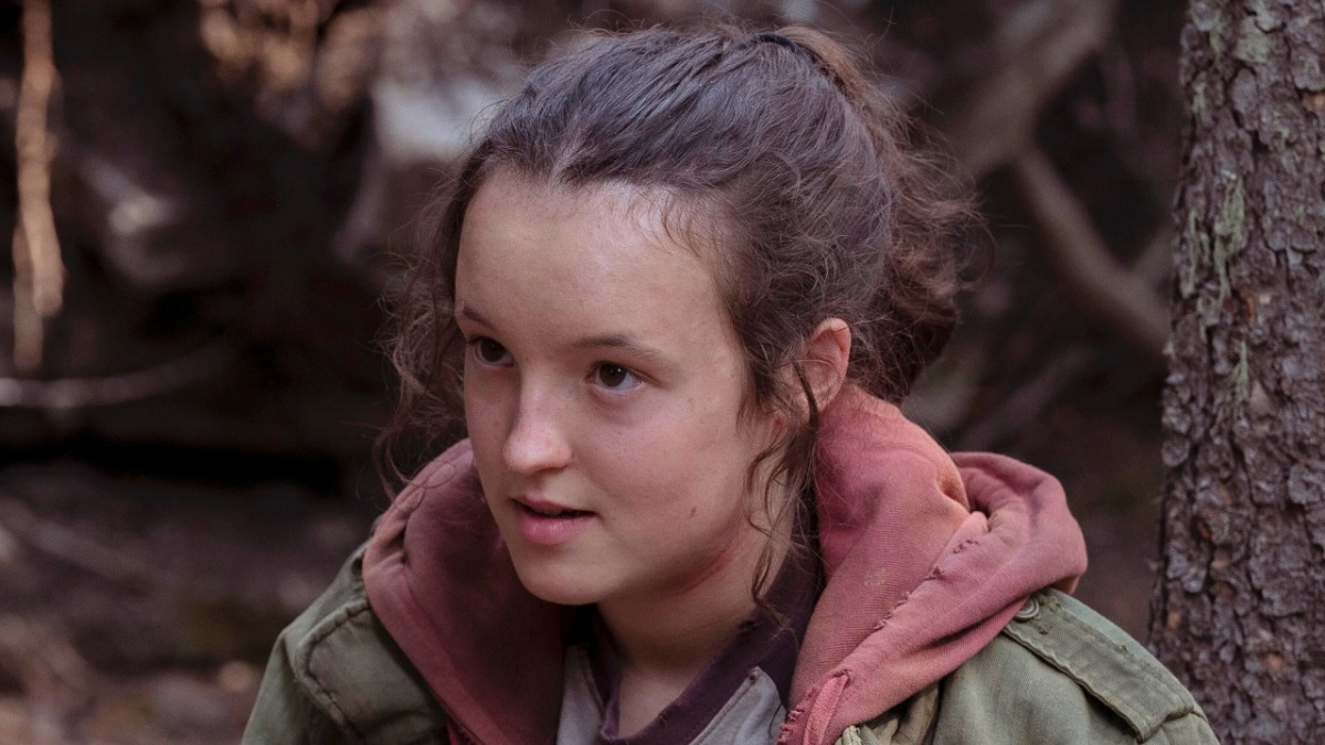 Image of Bella Ramsey as Ellie on HBO's 'The Last of Us.' Ellie is a white teen girl with her long brown hair in a ponytail wearing a grey and maroon jersey and red hoodie under a green jacket. She's sitting against a tree in the woods.
