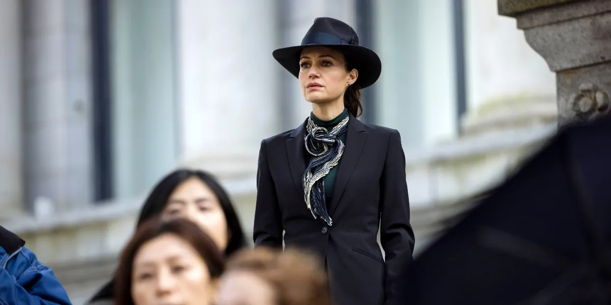 A woman (Carla Gugino) stands in above the crowd wearing black hat and trench coat in 'The Fall of the House of Usher.'
