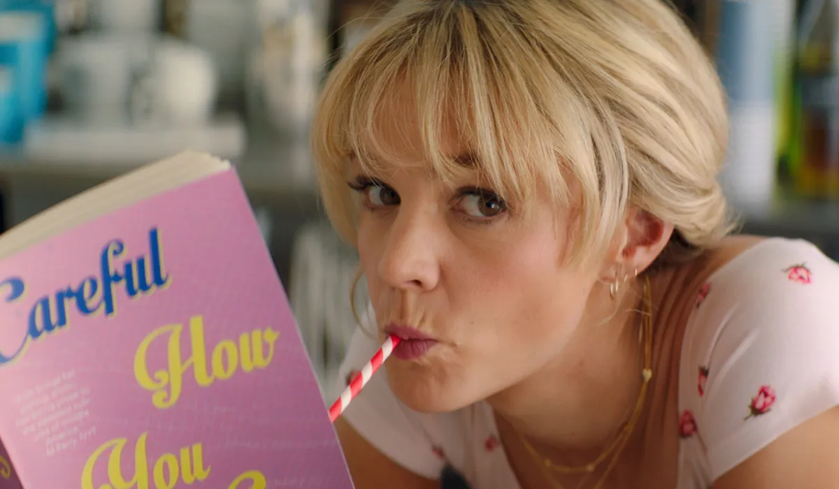 Carey Mulligan in Promising Young Woman, reading a book and drinking from a straw. 