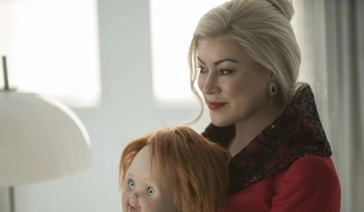 A blonde woman (Jennifer Tilly) wears a red coat and holds the Chucky doll in her lap in 'Cult of Chucky'.