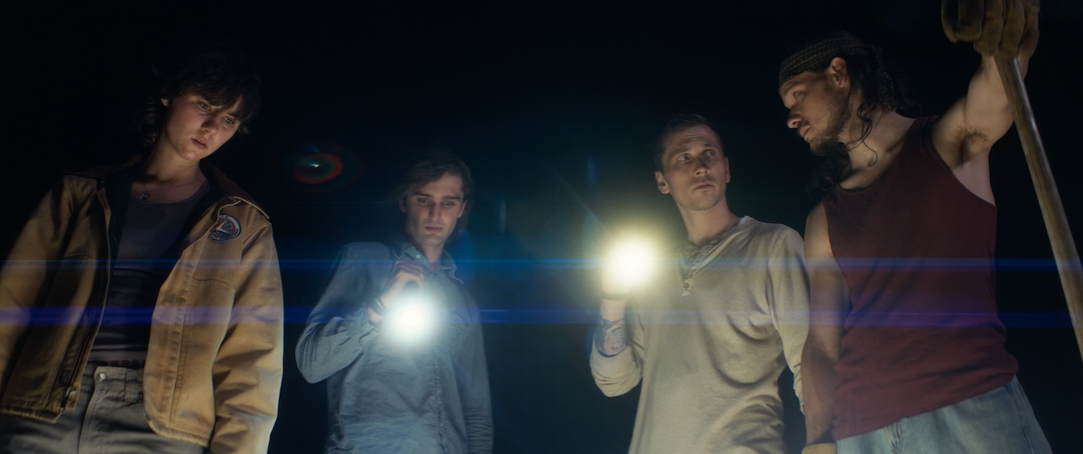 Four young people look down at the ground with flashlights against a black sky.
