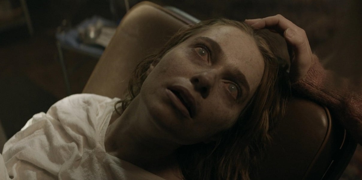 Image of Faustine Koziel as Lily on AMC's 'Daryl Dixon.' She is a white woman with long, red hair wearing a white hospital gown. She's lying on a gurney with the expression and clouded-over eyes of someone who's just become a zombie. 