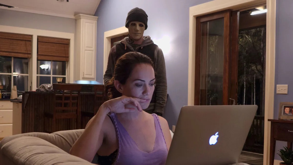 A masked man sneaks up behind a woman operating a laptop "It becomes quiet"