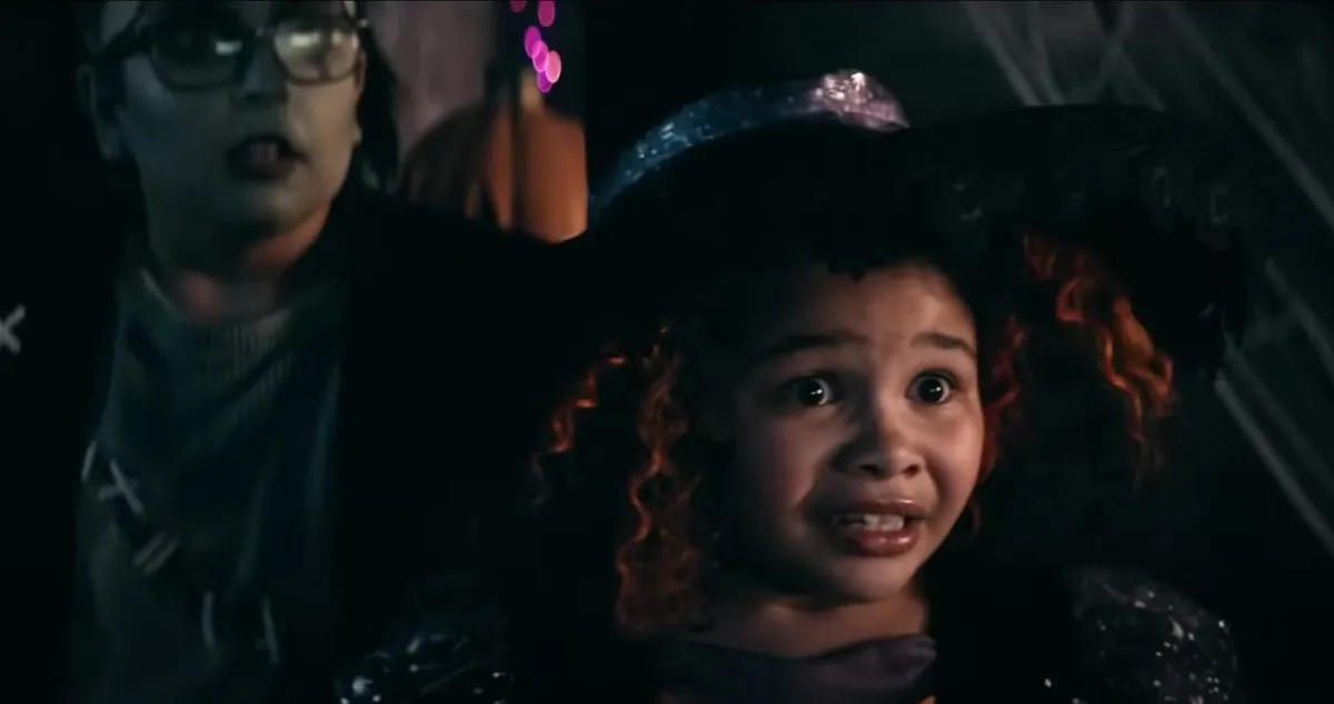 Screencap from the Jack in the Box short film, 'Feeding Time.' In the foreground is an elementary school age Black Girl dressed as a witch for Halloween. She has curly, shoulder length red hair and has a frightened expression on her face. Behind her is a little white boy in glasses dressed like Frankenstein's monster, also looking scared.