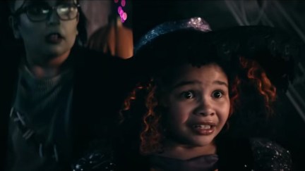 Screencap from the Jack in the Box short film, 'Feeding Time.' In the foreground is an elementary school age Black Girl dressed as a witch for Halloween. She has curly, shoulder length red hair and has a frightened expression on her face. Behind her is a little white boy in glasses dressed like Frankenstein's monster, also looking scared.