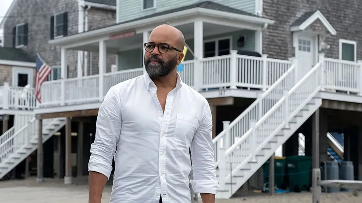 Image of Jeffrey Wright as Monk in the film 'American Fiction.' Monk is a light-skinned Black man who is bald with a salt-and-pepper beard. He's wearing black rimmed glasses and a white button down with the top buttons unbuttoned as he stands outside in front of a small town railroad station.