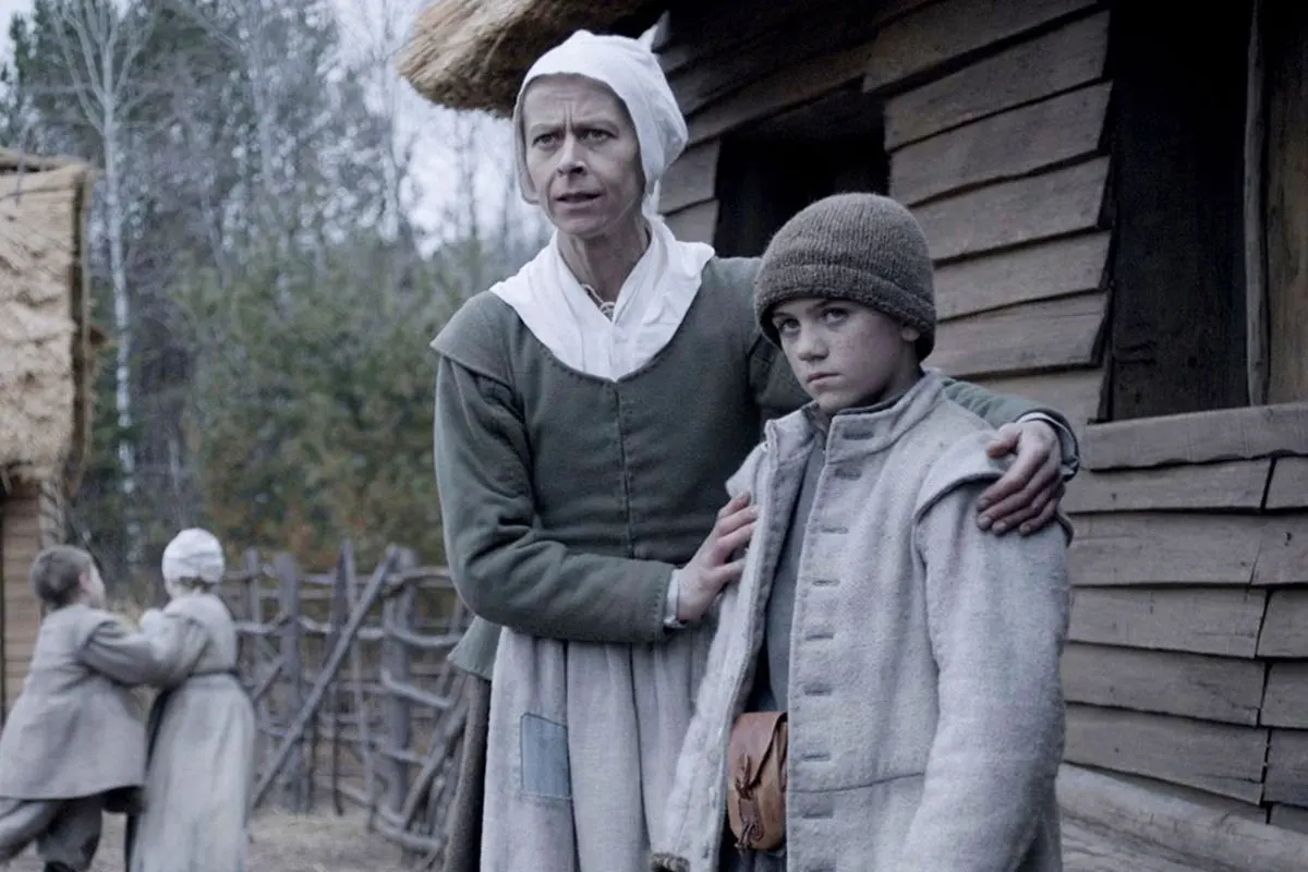 Kate Dickie as Katherine in The Witch, dressed in Puritan clothing, standing outside a cottage with Katherine's son Caleb 