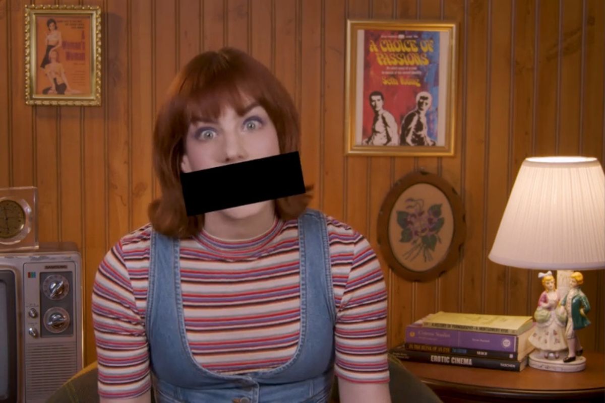 Maggie Mae Fish on a set in front of very '70s things like wood paneling, retro gaming station, and erotic books, posters, and more. She's being bleeped out while talking about sex for an 'Unrated' trailer.