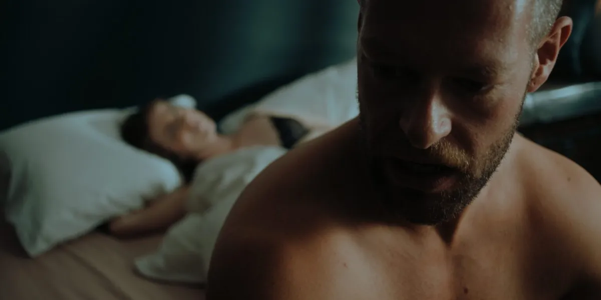 A post-coital Axel (Shane Quigley-Murphy) sits in the foreground with an undressed Dakota (Paige Gilmour) in the background in ‘Kill Your Lover.’