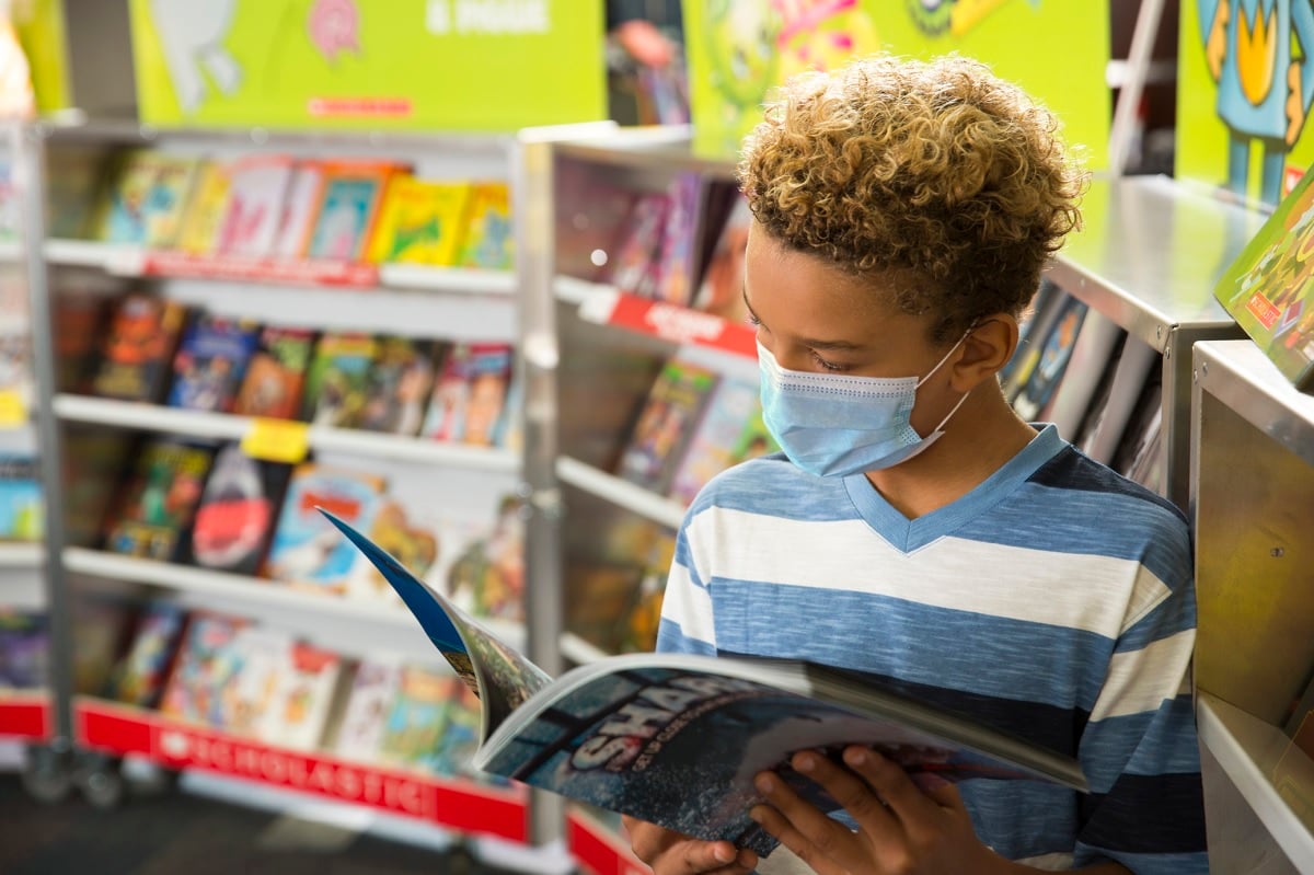 A child with a face mask on reading at a Scholastic book fair, with shelves of books out of focus in the background.