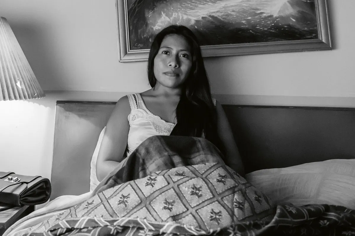 Black and white image of Yalitza Aparicio in the film 'Roma.' She is an Indigenous Mexican woman with long, black hair. She's sitting up in bed wearing a white nightgown with her blankets pulled up over her knees. A painting on the wall behind her is crooked, and her purse sits on the nightstand behind her.