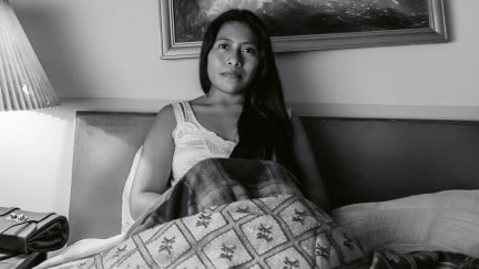 Black and white image of Yalitza Aparicio in the film 'Roma.' She is an Indigenous Mexican woman with long, black hair. She's sitting up in bed wearing a white nightgown with her blankets pulled up over her knees. A painting on the wall behind her is crooked, and her purse sits on the nightstand behind her.