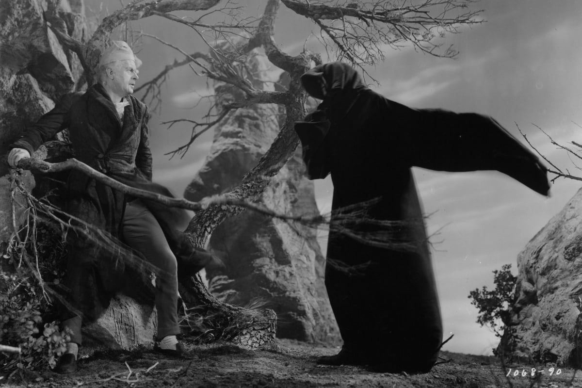 Still from the 1938 A Christmas Carol; a black and white image of Reginald Owen as Scrooge, wearing bed clothes and leaning back in horror from a grim reaper type figure in a graveyard