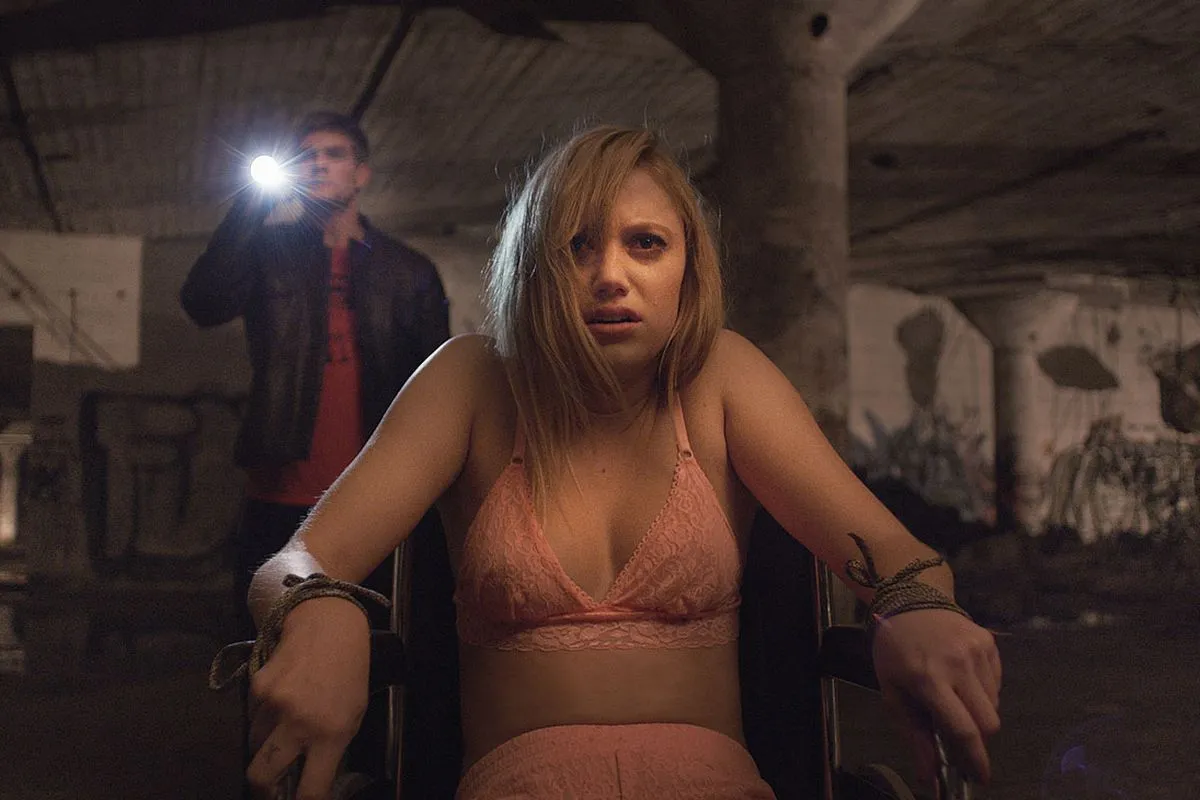 A girl tied to a chair looks at something offscreen in horror as a boy with a flashlight stands behind her in "It Follows"