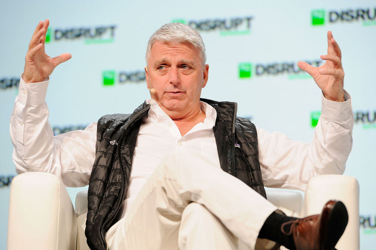 A middle aged white guy (John Riccitiello) wears a puffy vest and gestures broadly while speaking during a conference.