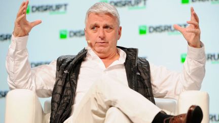A middle aged white guy (John Riccitiello) wears a puffy vest and gestures broadly while speaking during a conference.