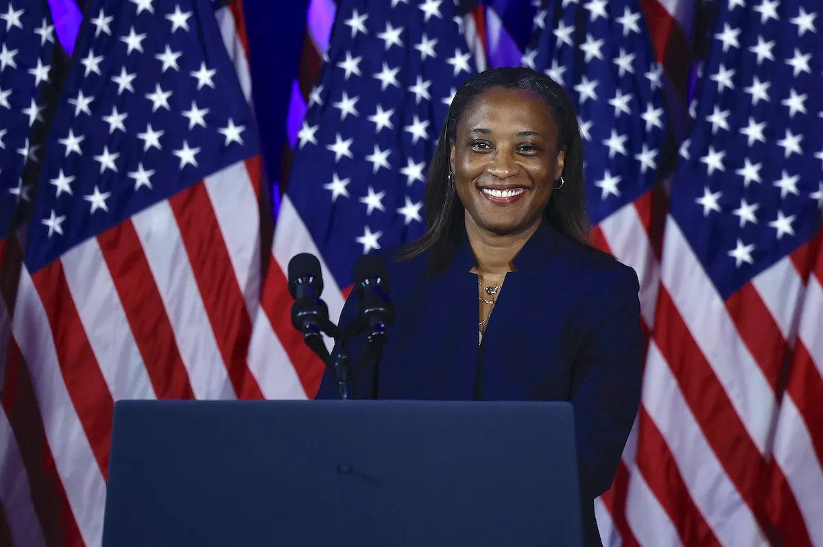 A Black woman (Laphonza Butler) stands at a podium smiling in front of American flags.