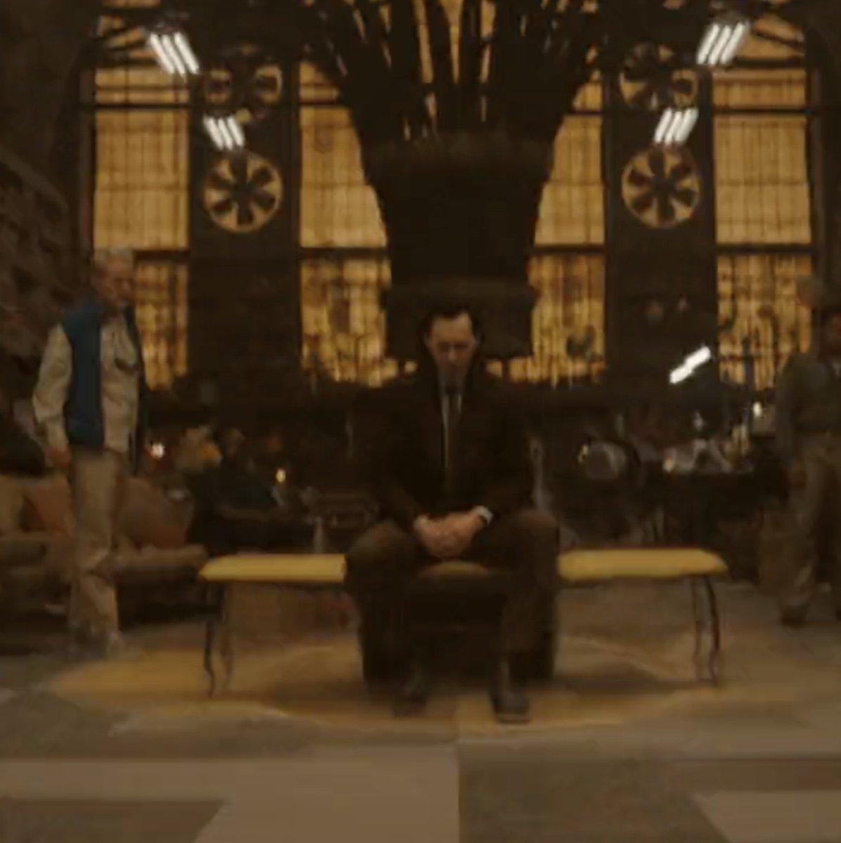 Blurry shot of Loki sitting in an early version of O.B.'s workroom. Mobius is visible next to him, wearing a white shirt and blue vest.