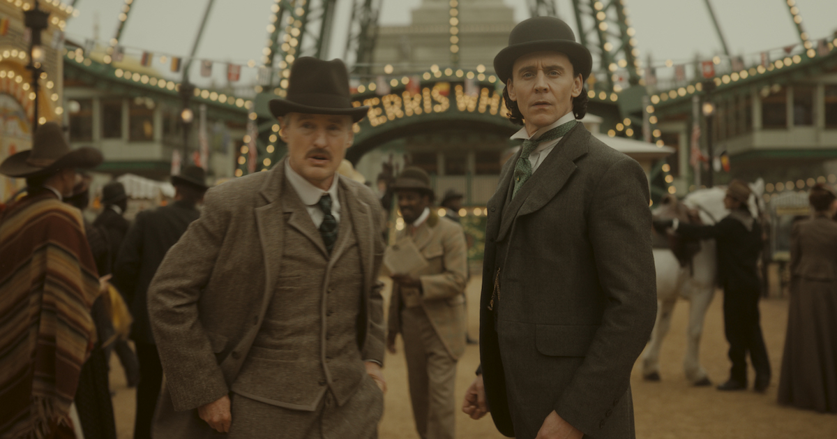 Mobius and Loki standing in front of the ferris wheel at the 1893 World's Fair. They're both in period costume.