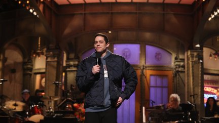 Pete Davidson stands on the SNL stage, holding a microphone.