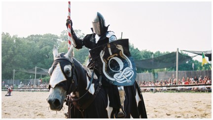 Stock photo of a jousting knight in armor, riding a horse as a crowd watches.