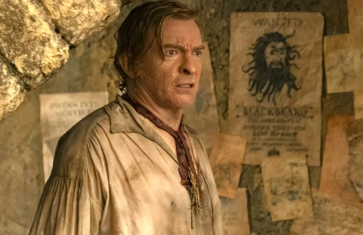 Rhys Darby as Stede Bonnet stands by a wanted poster of Blackbeard in 'Our Flag Means Death' season 2