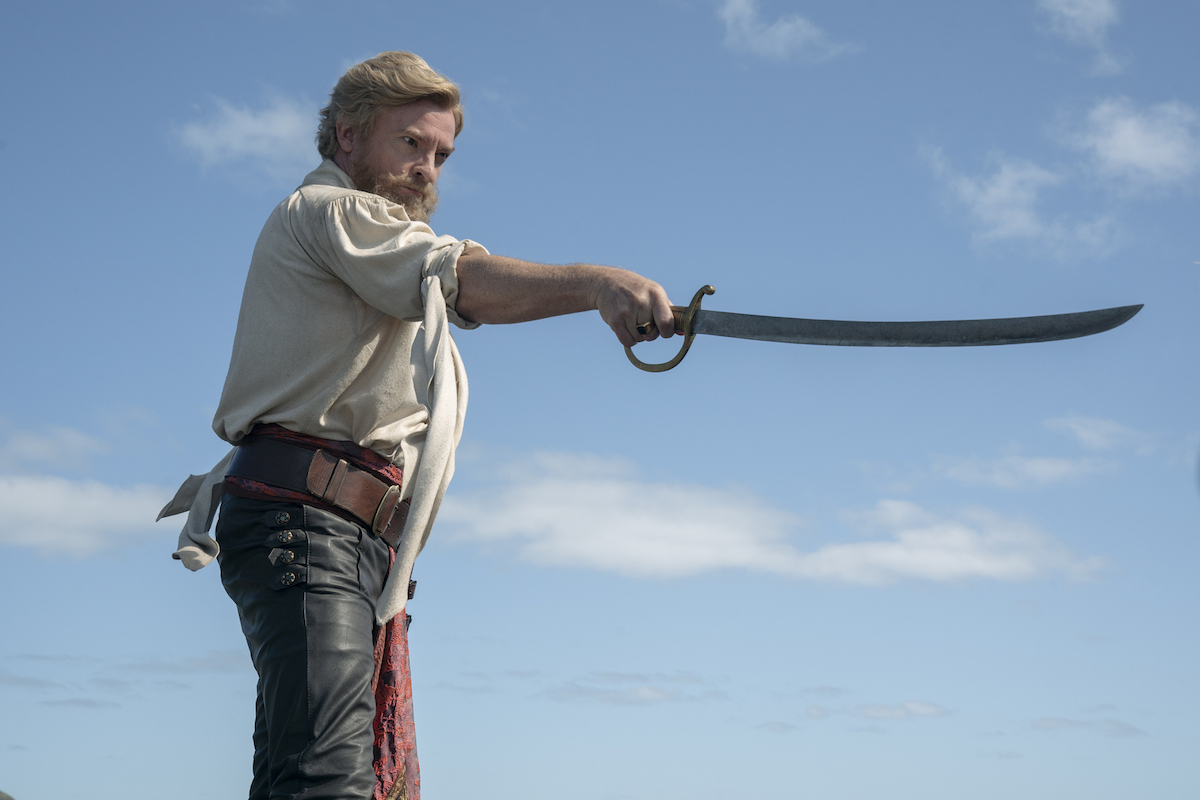 Stede holds a sword out against a backdrop of a blue sky. He has a beard.