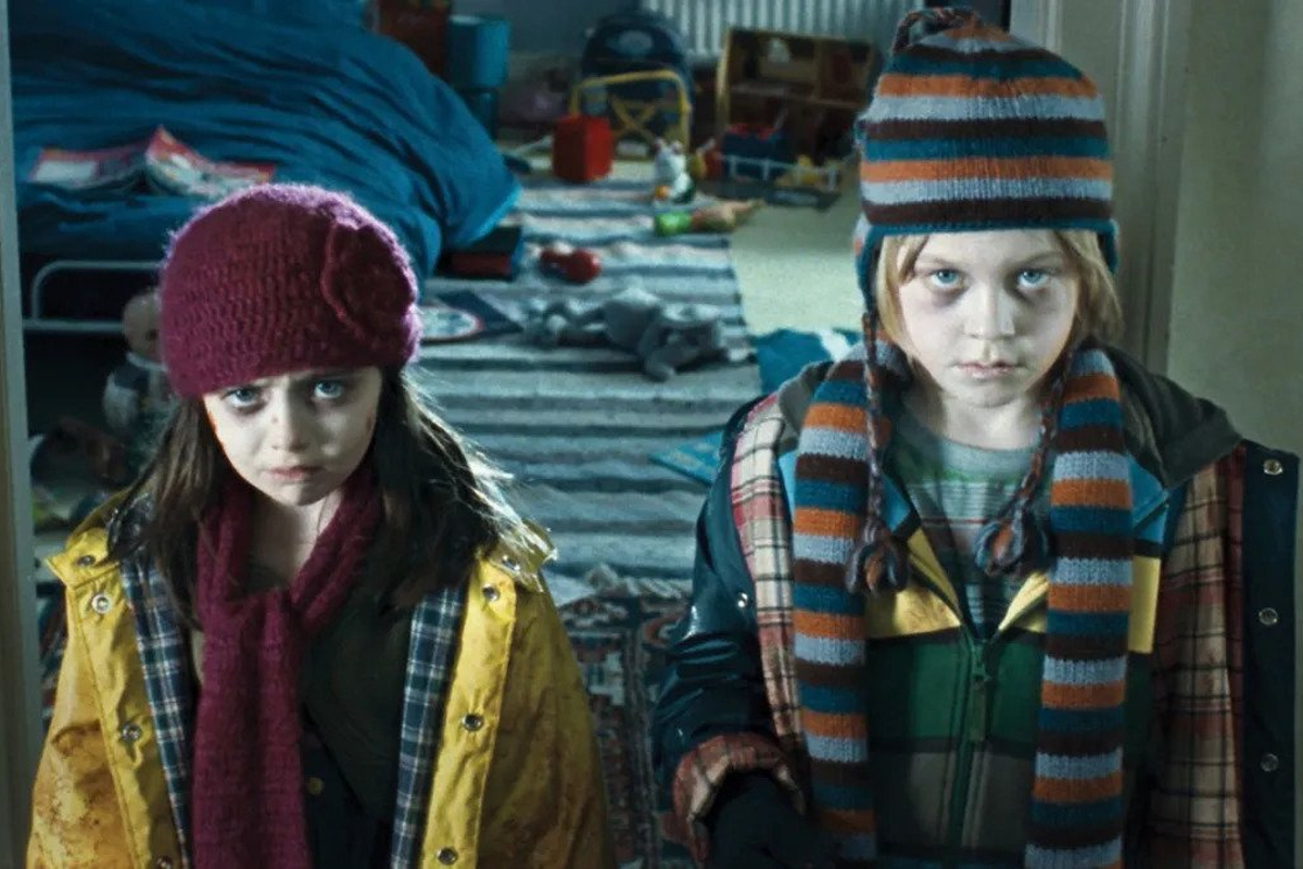 Still from The Children; two white children, a dark haired girl and red haired boy, stare into the camera. They're wearing outdoor winter wear and have dark circles under their eyes.