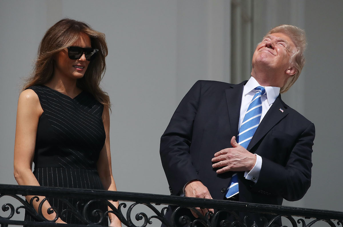 Donald Trump stares at the sky, squinting, as Melania stands next to him wearing dark sunglasses.