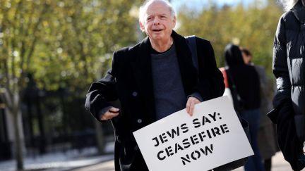 Wallace Shawn walks down the street, holding a sign that says 