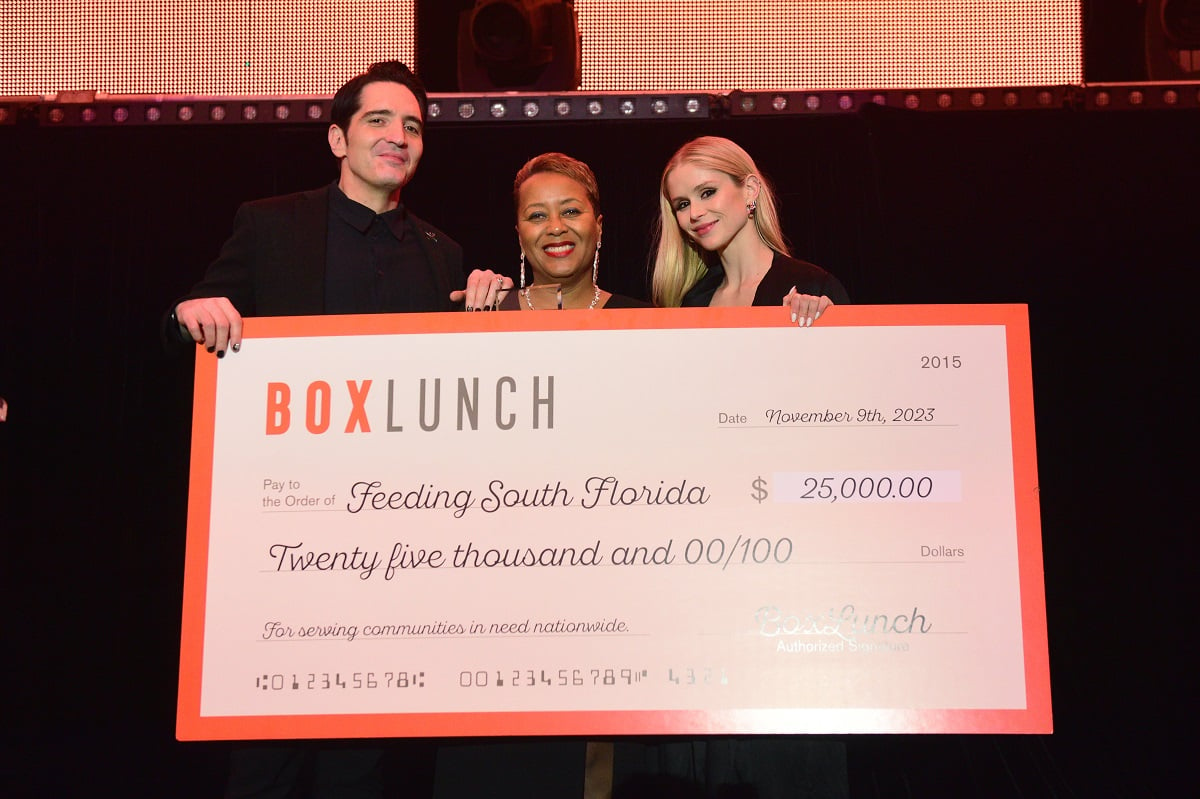 Image of David Dastmalchian, Allyson Vaulx of Feeding South Florida, and Erin Moriarty standing together on stage holding a large fake $25,000 check from BoxLunch to Feeding South Florida. David is a white man with short black hair wearing an all-black suit. Allyson is a Black woman with short red hair wearing a black dress and a diamond necklace and earrings, and Erin is a white woman with long blonde hair wearing a black dress. 
