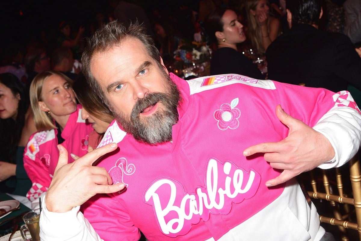 Image of David Harbour seated at a table in a crowded ballroom. He is a white man with brown hair and a salt-and-pepper beard. He's wearing a pink and white BoxLunch 'Barbie' racing jacket and making a silly face as he holds up his hands like he's rocking out.