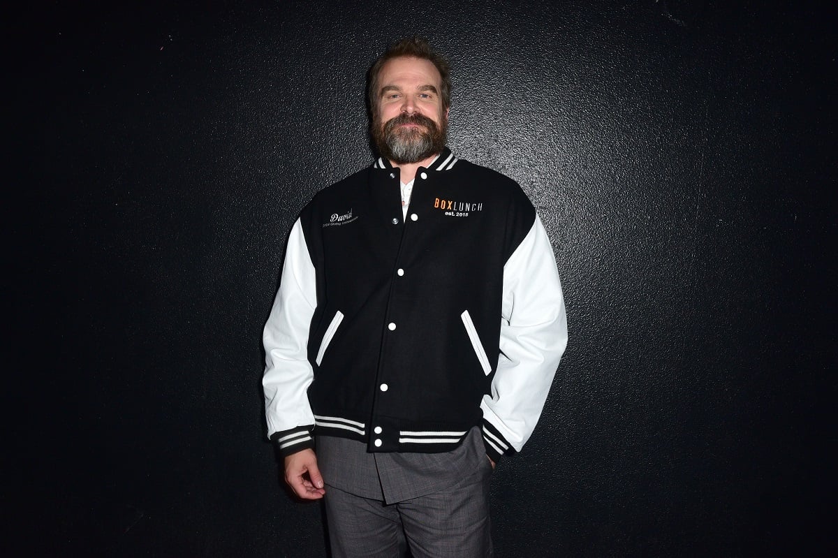 Image of David Harbour in front of a black wall. He is a white man with brown hair and a salt-and-pepper beard. He's wearing a black and white BoxLunch letterman jacket and grey suit pants and smiling at the camera with one hand in his pocket.
