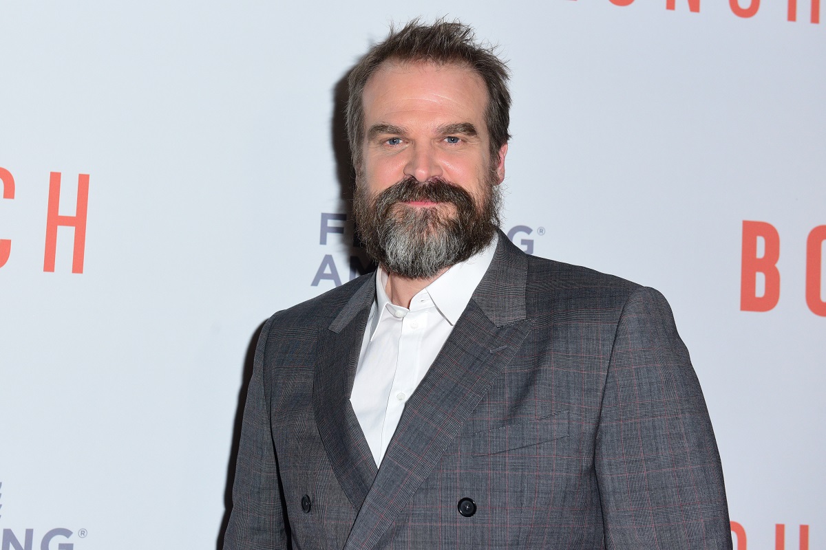 Image of David Harbour in front of a step and repeat with the logos for BoxLunch and Feeding America on it. He is a white man with brown hair and a salt-and-pepper beard. He's wearing a grey suit with a white buttondown and smiling at the camera.