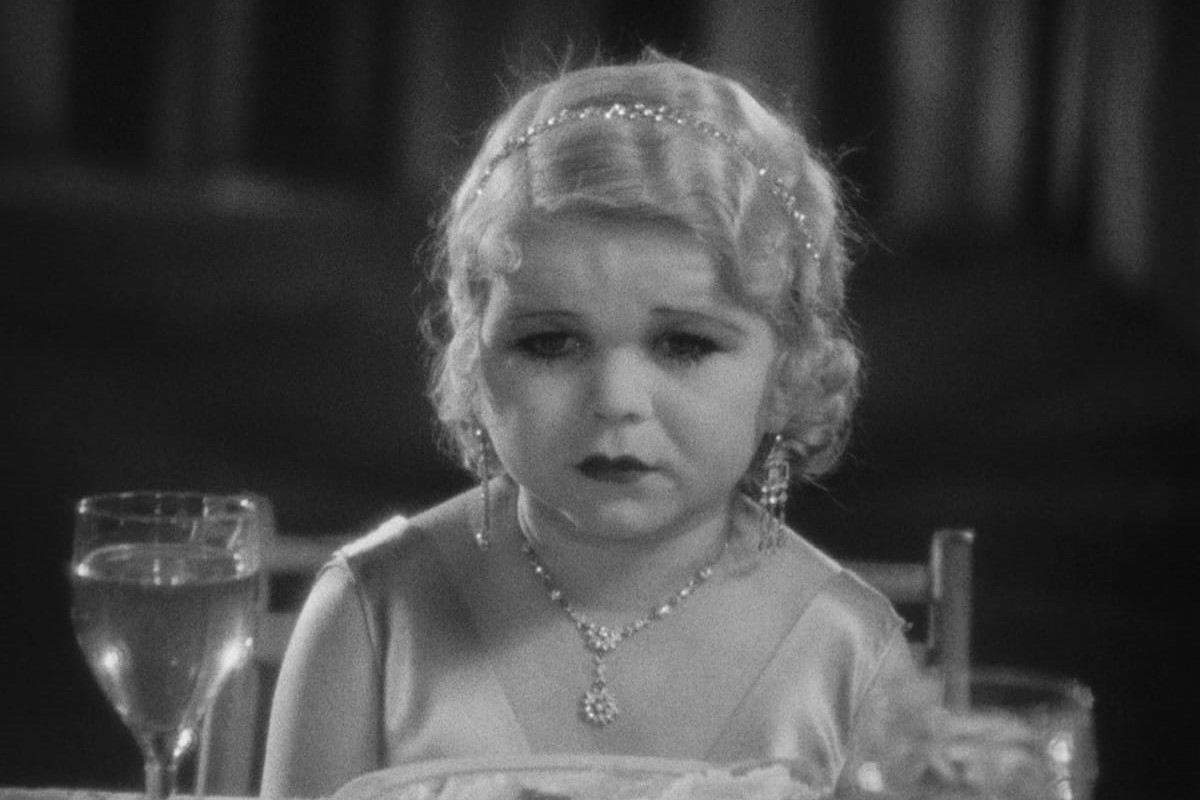 Black and white image of Daisy Earles (Frieda) in a scene from 'Freaks.' She is a blonde, white woman with dwarfism wearing a thin, sparkly tiara, dangling earrings, and a sparkly necklace with a white satin sleeveless dress. She's seated at a dinner table and looking off into the distance with a sad expression.