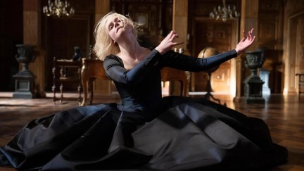 Image of Elle Fanning as Catherine in a scene from Hulu's 'The Great.' She is a white woman with chin-length straight blonde hair wearing an 18th century black dress. She is kneeling on the floor of an ornate office in her palace mid-dance with her arms extended to her left.