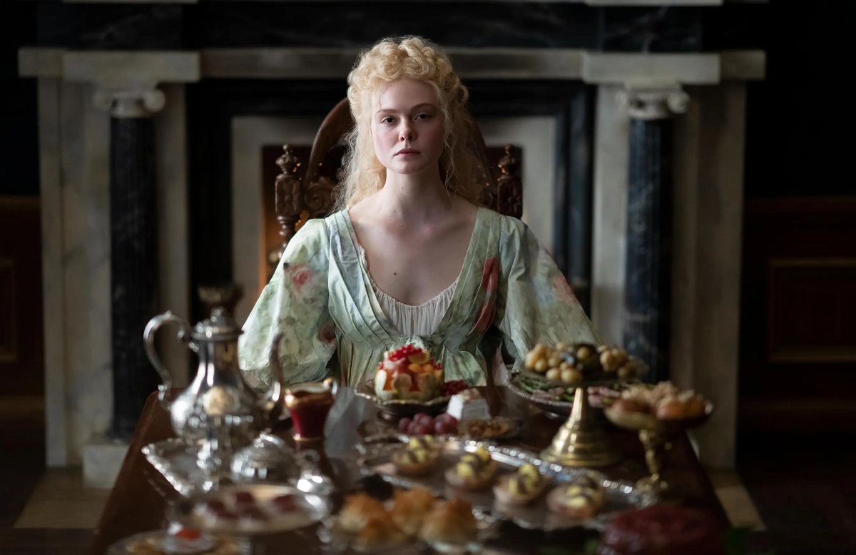Image of Elle Fanning as Catherine in a scene from Hulu's 'The Great.' She is a white woman with long, wavy blonde hair wearing an 18th century green floral robe and white nightgown. She is sitting at the end of a long dining table with food spread out before her. She's wearing a serious expression. 