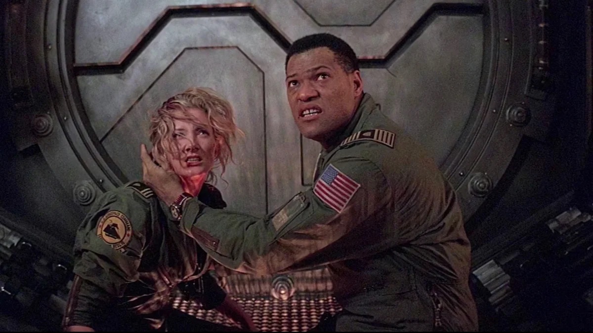 Joely Richardson (Starck) and Lawrence Fishburne (Miller) in a scene from 'Event Horizon.' Starck is a white woman with blonde wavy hair wearing a green jumpsuit uniform. Miller is a Black man with closely cropped black hair and also wearing a green jumpsuit uniform. He is partly shielding her face as they both look up at something frightening. 