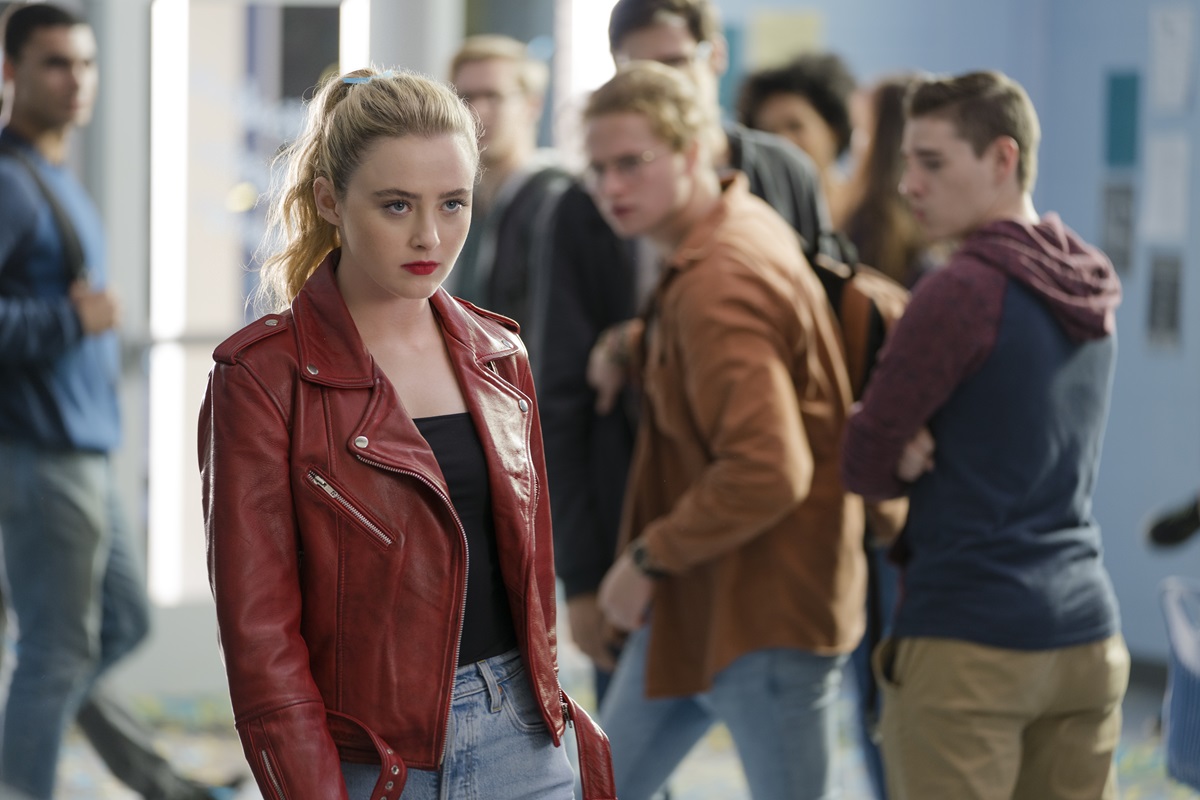 determined looking teenager in red leather jacket walks through school halls as students look on