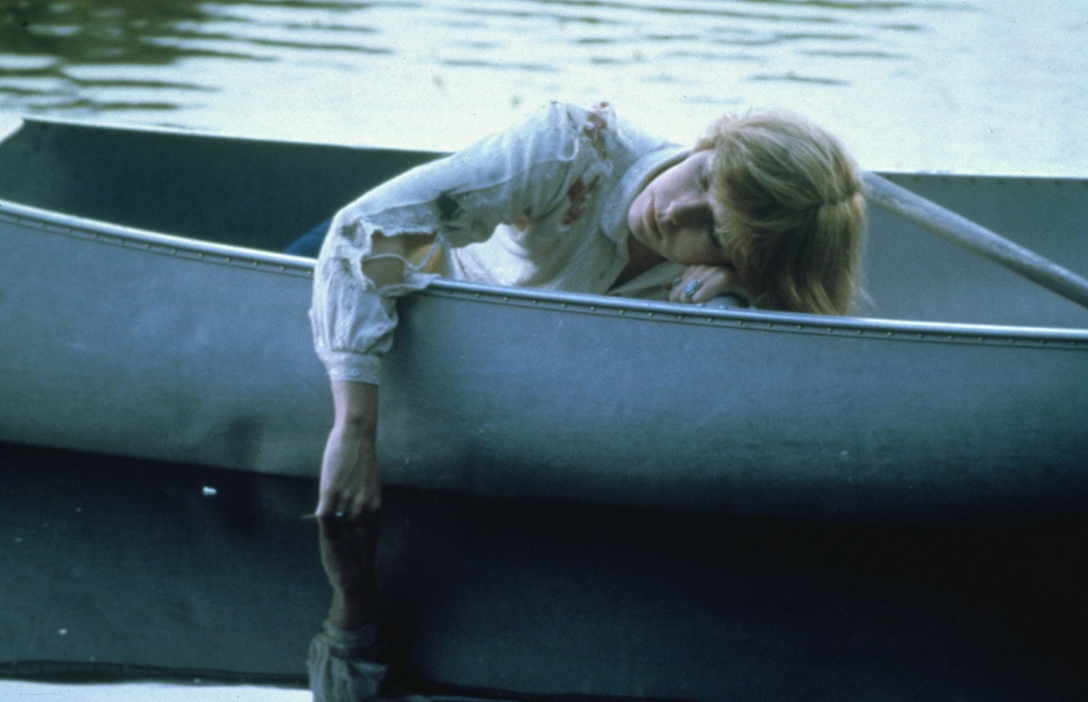 Adrienne King as Alice in a scene from 'Friday the 13th.' She is a white teen girl with short red hair wearing a long-sleeved white shirt that's been torn in a fight. She's sleeping in a canoe out on a lake with her arm dangling in the water. 
