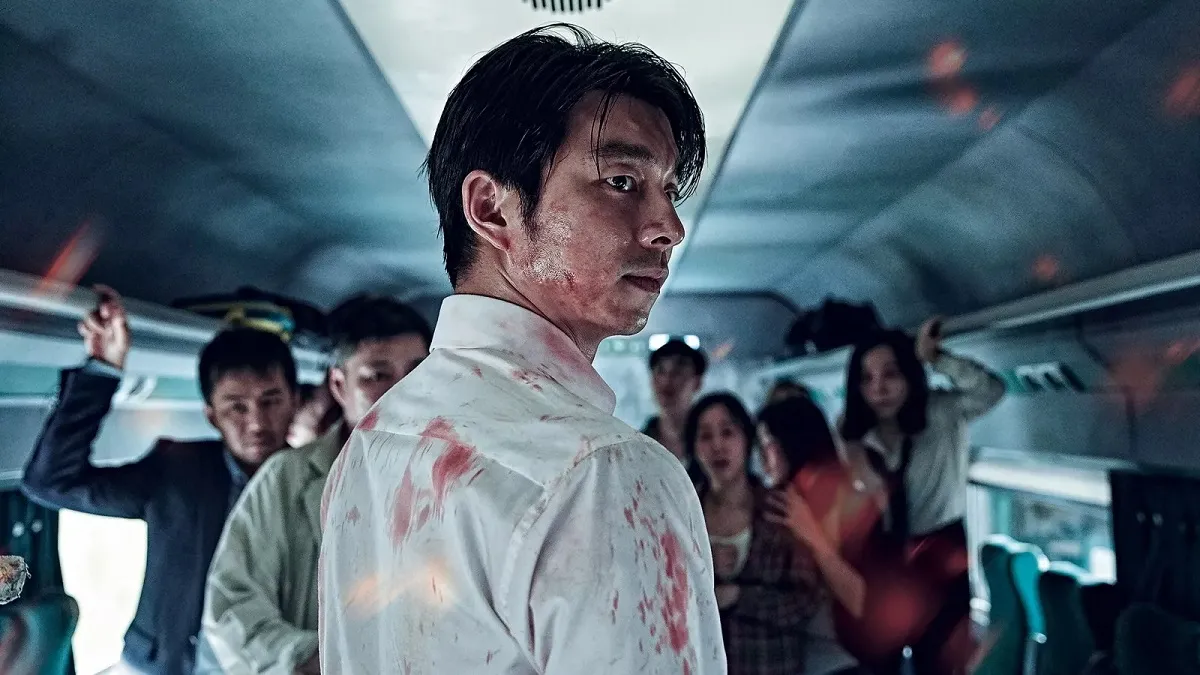 Gong Yoo in a scene from the fllm 'Train to Busan." Yoo is a Korean man with dark hair that hangs in his eyes. He's wearing a white buttondown that is splattered with blood. His face is also sweaty and splattered with blood as he seriously looks over his shoulder while standing on a train with other frightened Koreans.