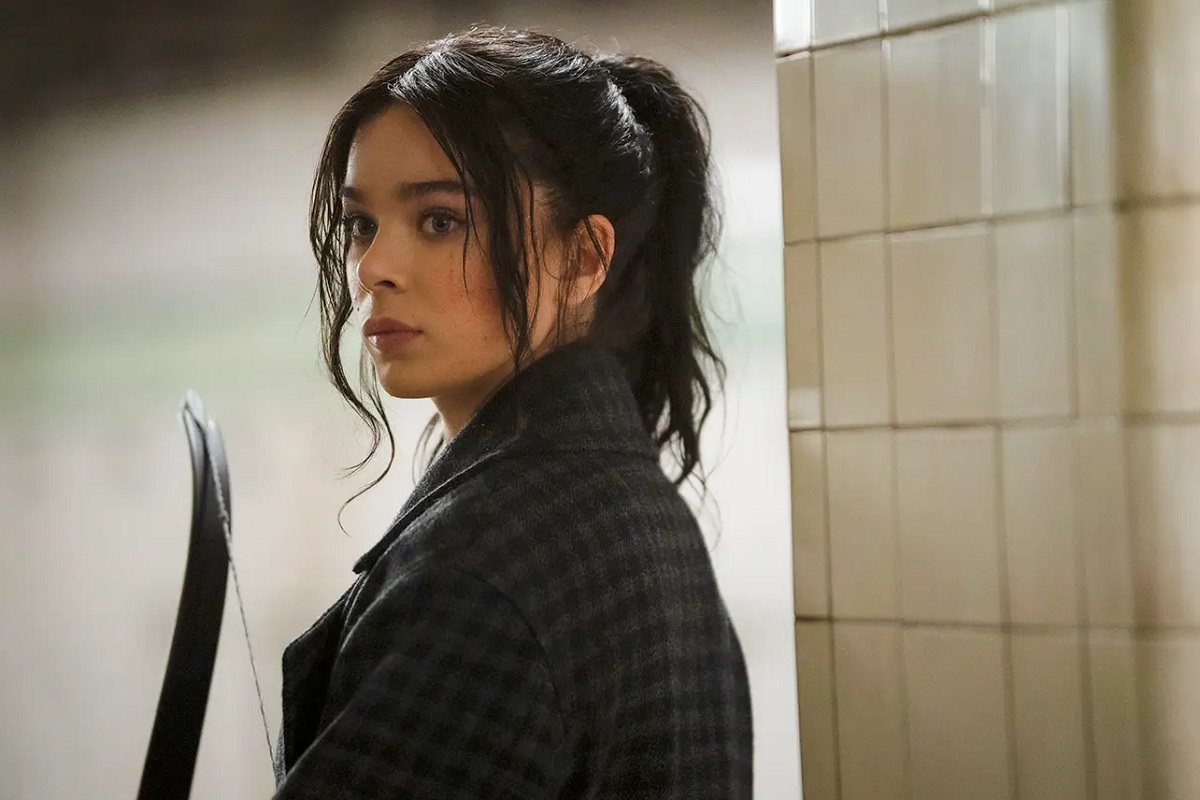 Image of Hailee Steinfeld as Kate Bishop in Disney+'s 'Hawkeye.' She is a white young woman with her long dark hair in a ponytail as shorter strands hang in her face. She's wearing a black gingham coat and holding a bow as she stands in a subway station looking over her shoulder. 