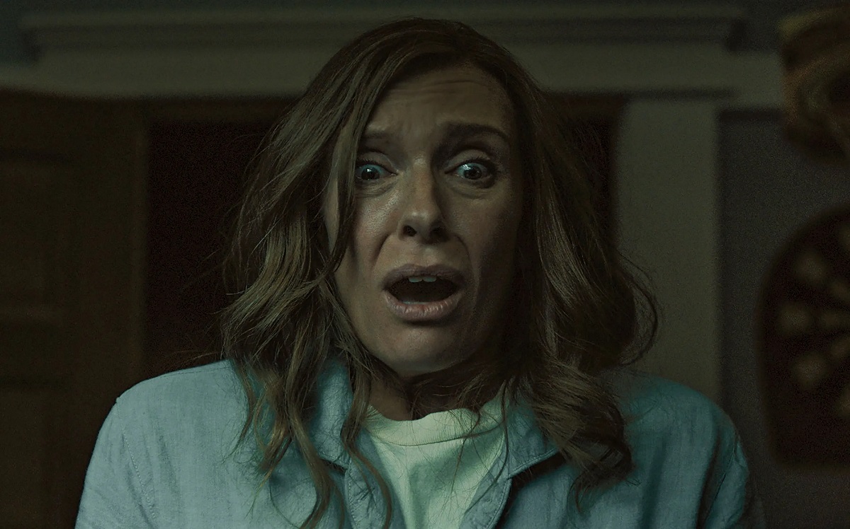 Toni Collette as Annie in 'Hereditary.' Annie is a white woman with shoulder-length strawberry blonde hair wearing a light blue buttondown over a white shirt. She is looking at something with a horrified expression on her face.
